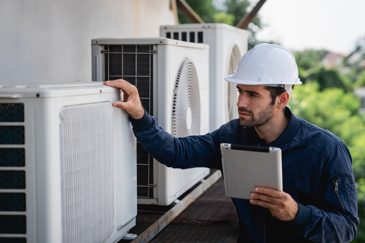 Optimal HVAC Performance: A maintenance worker in a hard hat inspects an outdoor air conditioning unit while holding a tablet.